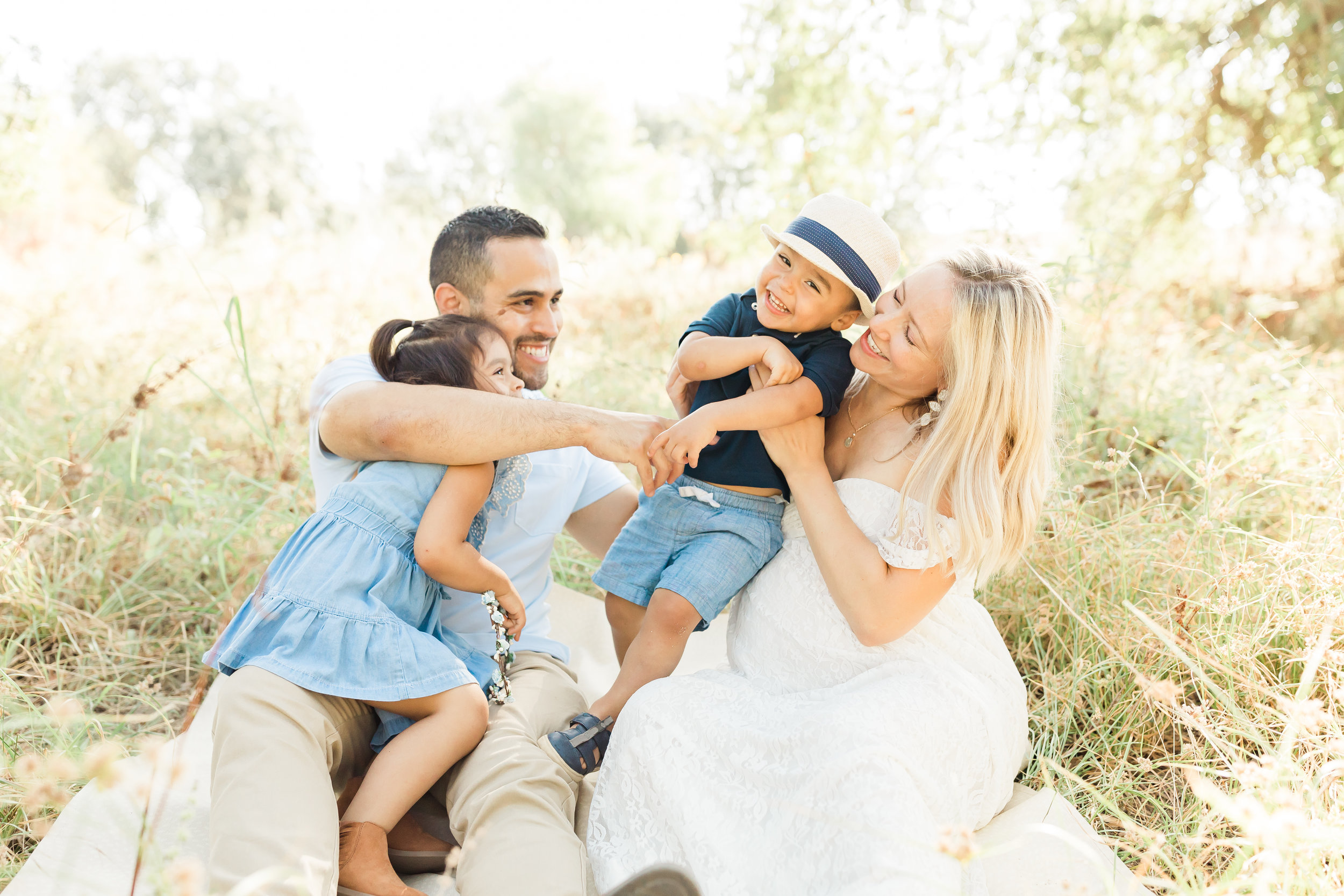 Family wearing neutrals and chambray sitting in a field tickling toddler boy. Maternity photos.