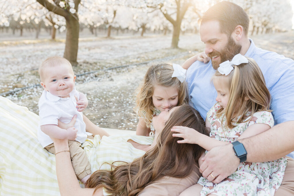family photo of family of 5 in almond blossom orchard. mom is laying on dad's lap with baby sitting on her lap and two little girls snuggle in. Positive mindset for family photos