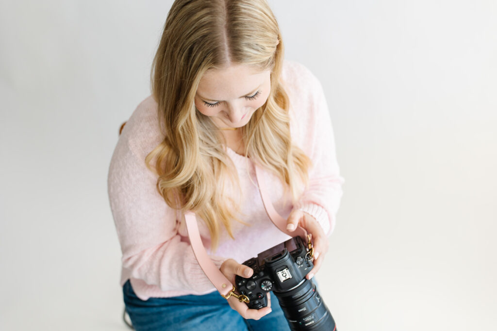 blonde haired new photographer female in pink sweater is seated in front of a white backdrop looking down at a dslr camera.